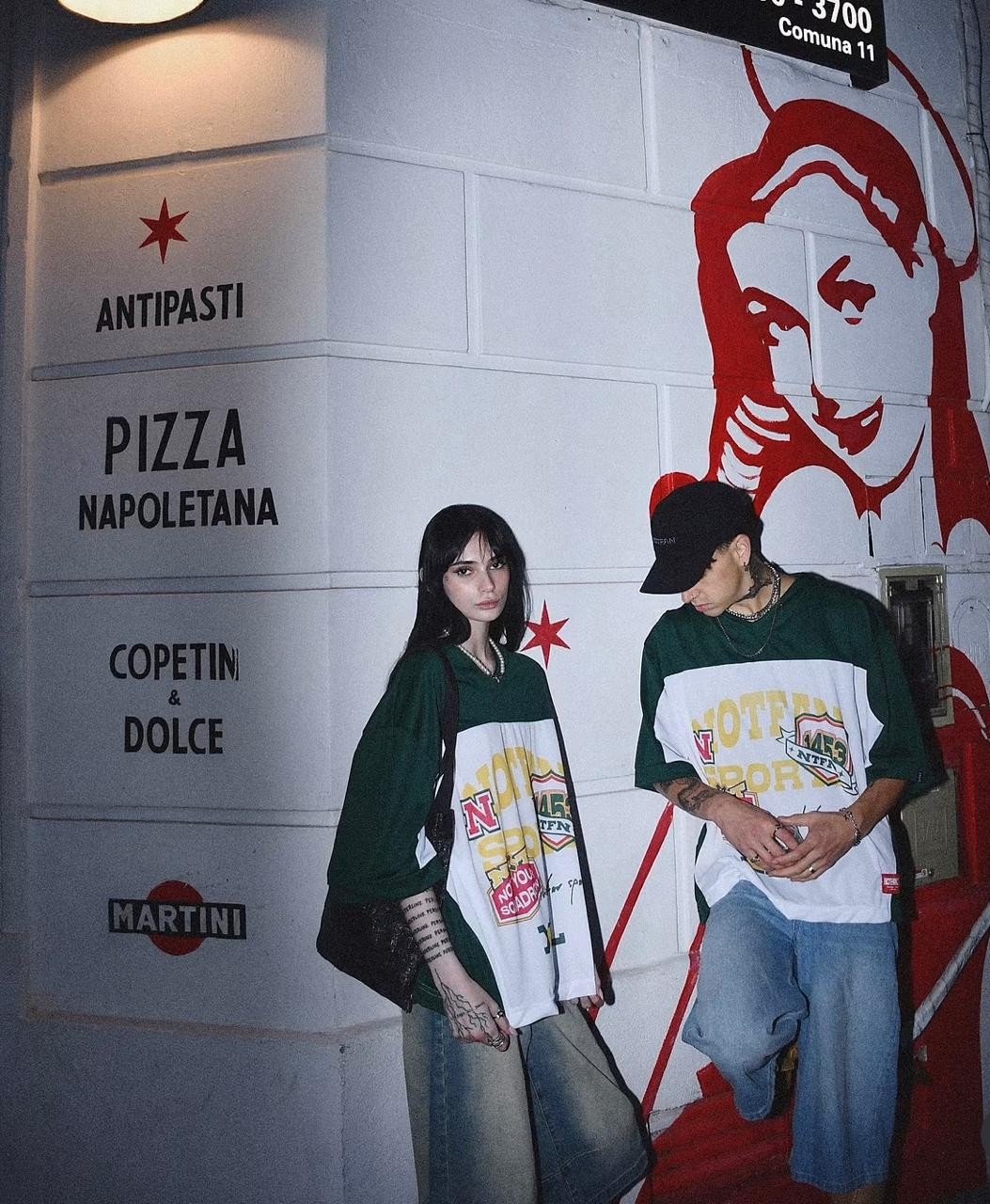 Dos modelos con la camiseta de fútbol americano verde y blanca frente a una pared con mural rojo