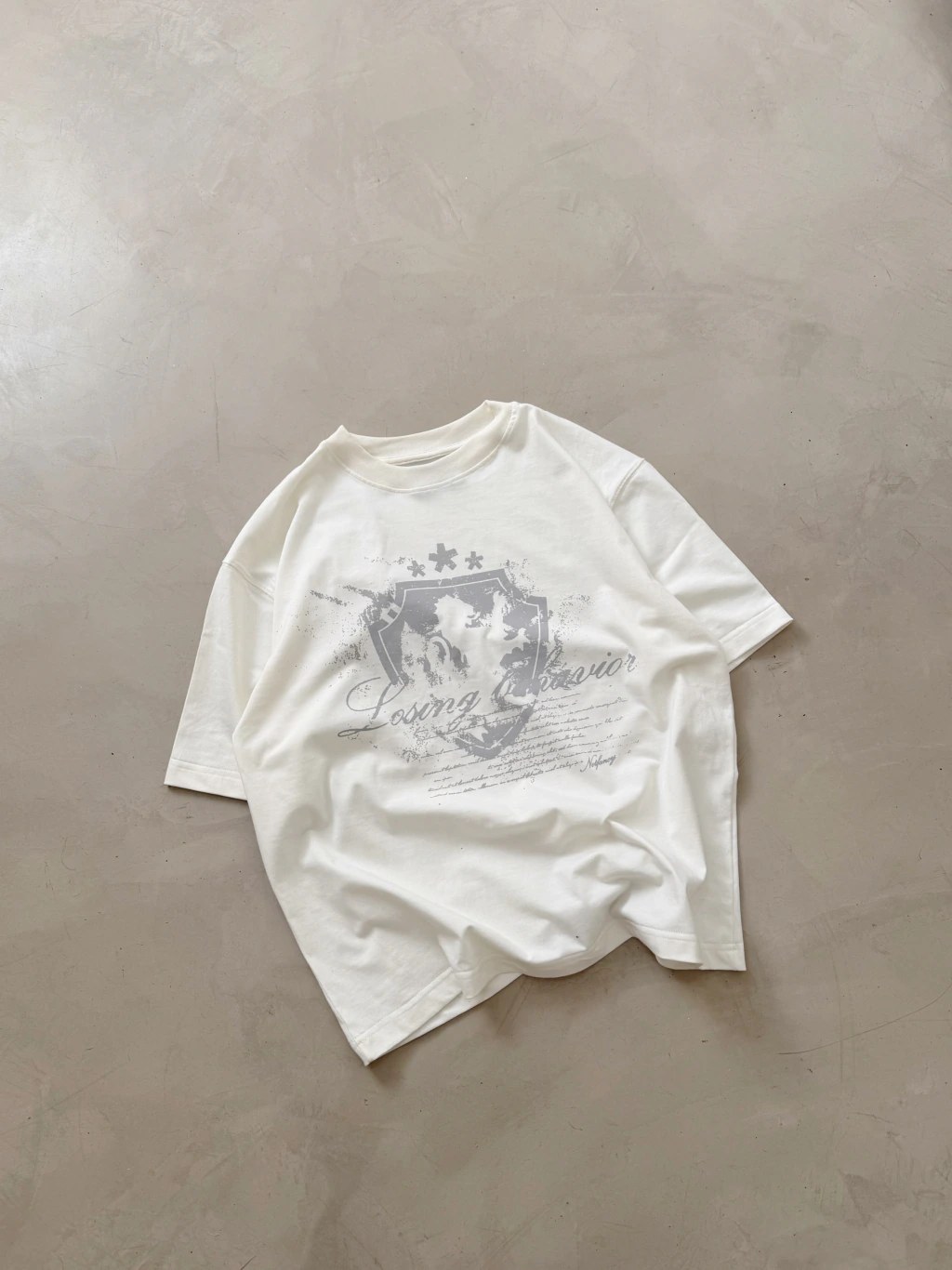 Remera blanca con la gráfica gris Losing Behavior sobre un piso de cemento